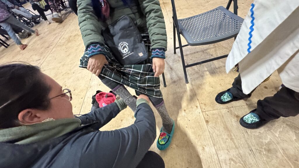 A woman bends down to help another person adjust their socks and brightly colored moccasins. The person seated wears a green jacket and a plaid skirt, while people in the background observe and chat.