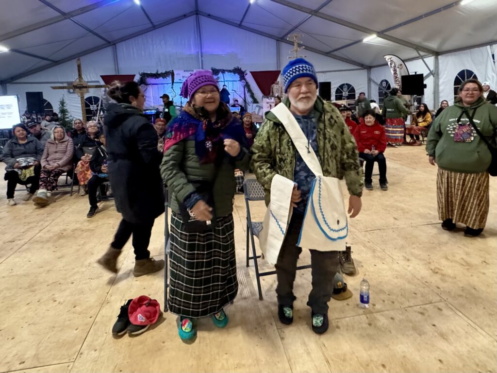 A Cree couple, Johnny and Louisa B. Saganash, stand side by side in the gathering tent, dressed warmly. The man wears a blue knitted hat and a large white sash over his green jacket, while the woman dons a purple hat and plaid skirt. They smile as they prepare for a traditional dance, surrounded by seated spectators.