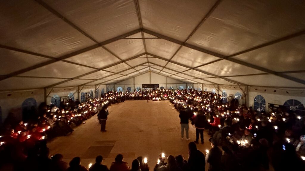 (Candlelight vigil in large tent): A wide view of a large gathering inside a tent, with rows of people seated in a circular formation around the perimeter. Everyone is holding a lit candle, illuminating the entire space in soft light. The atmosphere is solemn as they participate in a candlelight vigil.