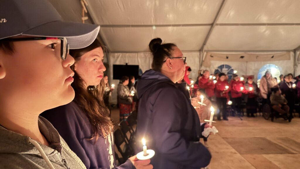 (Candlelight vigil attendees close-up): A close-up of several individuals standing and holding lit candles, their faces illuminated by the warm candlelight. The expressions on their faces are reflective and focused, showing the emotional weight of the moment during the vigil.