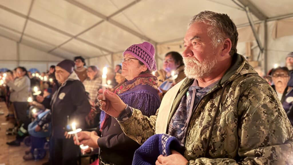 (Johnny and Louisa B. Saganash holding candles): A close-up of Johnny and Louisa B. Saganash, an elder Cree couple, holding candles during the vigil. Johnny wears a camouflage jacket, and Louisa wears a purple knit hat and a scarf, both looking solemn and thoughtful.