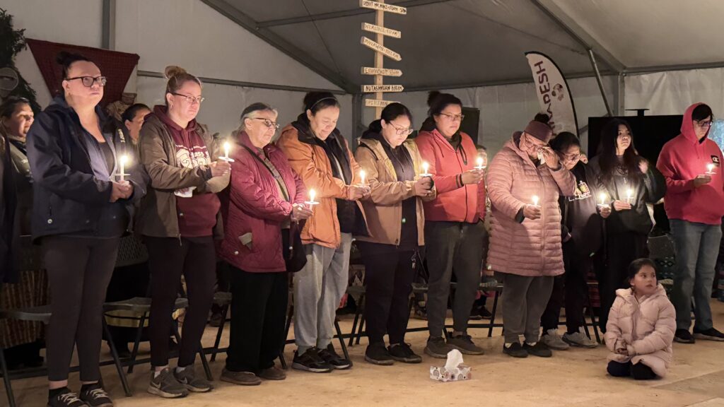 (Guanish family members during the vigil): A group of women and one young girl, all standing side by side and holding lit candles during the vigil. One woman wipes her eyes as others stand with their heads bowed, reflecting on the loss of George Amos Guanish. The atmosphere is filled with quiet support and comfort.