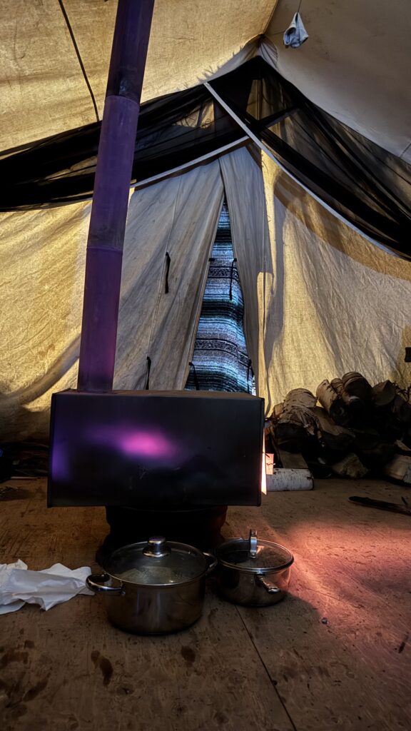 Inside a Naskapi tent, a metal stove is glowing with purple light from a fire within. Two large cooking pots sit on the ground in front of the stove, and birch logs are stacked to the side. The tent's interior is softly lit, with a blanket covering the entrance flap.