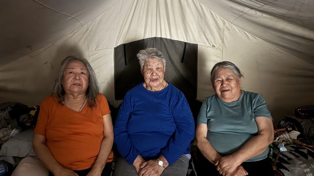 Three elderly Naskapi women sit together inside their tent, smiling gently at the camera. The one on the left wears an orange shirt, the middle elder wears a royal blue shirt, and the one on the right wears a light teal shirt. Behind them, the canvas of the tent walls forms a cozy backdrop. They are seated on a blanket-covered surface, sharing their presence and stories.