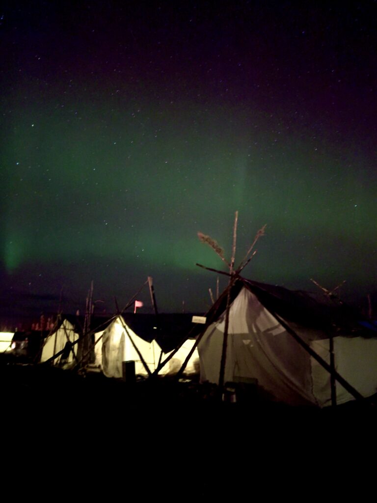 The Northern Lights (aurora borealis) shimmer across the night sky in green and purple hues. In the foreground, traditional Naskapi tents are visible, their canvas covers lit from within, with wooden supports silhouetted against the vibrant auroras.