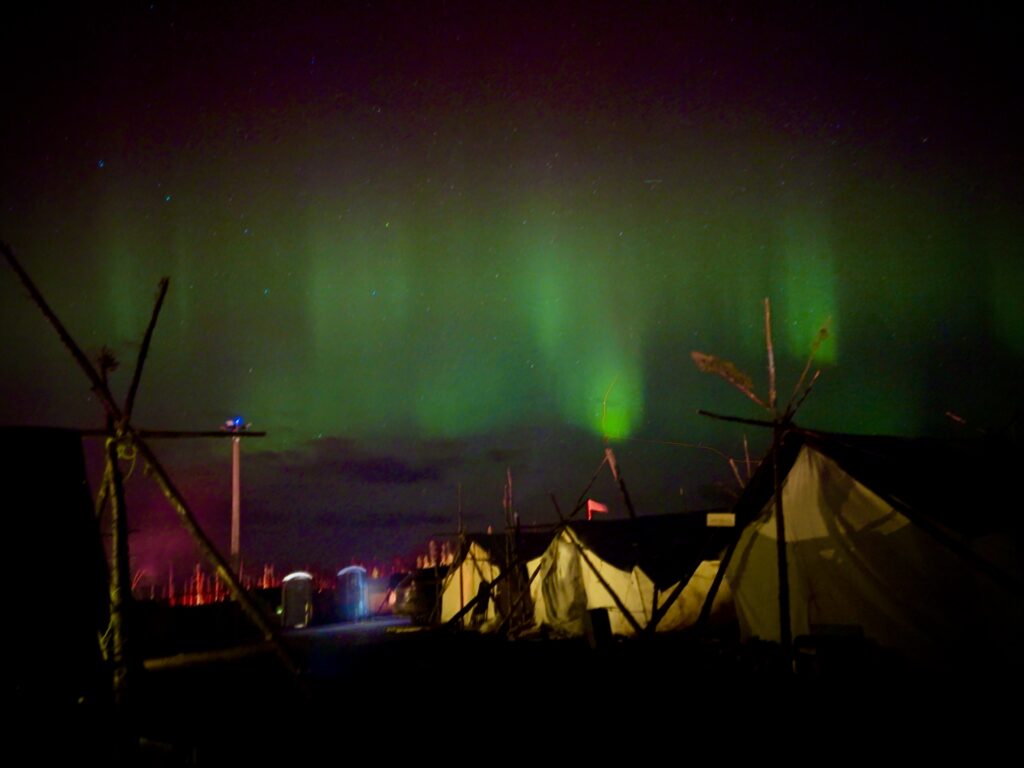 The aurora borealis glows brightly in shades of green, casting an ethereal light over the camp. Naskapi tents stand in the foreground, their outlines stark against the celestial display, while a few lights from the camp area dot the background.