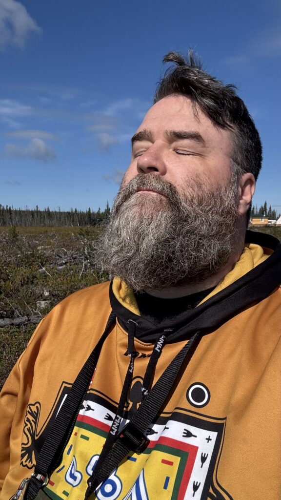 Benjamin Jancewicz stands outdoors, eyes closed and face lifted toward the sun, fully immersed in the warmth and light. He wears a brightly colored hoodie with intricate patterns, and the background shows an expanse of forest and clear blue sky, evoking a moment of deep connection to nature and tranquility.