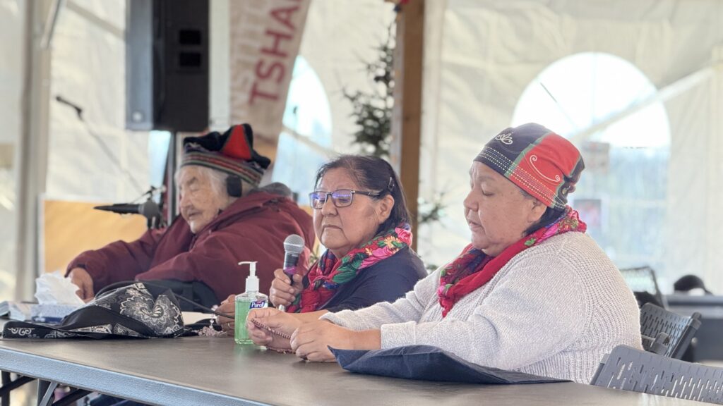 A panel of three Naskapi women sits at a table under a large tent. The woman in the center holds a microphone, sharing stories with those in attendance. She wears a floral scarf, a symbol of the traditional style, and the other women also don similar scarves. In the background, an older woman, dressed warmly, listens through headphones. On the table are a bottle of hand sanitizer and a black purse, contrasting with the serious atmosphere as the stories unfold.