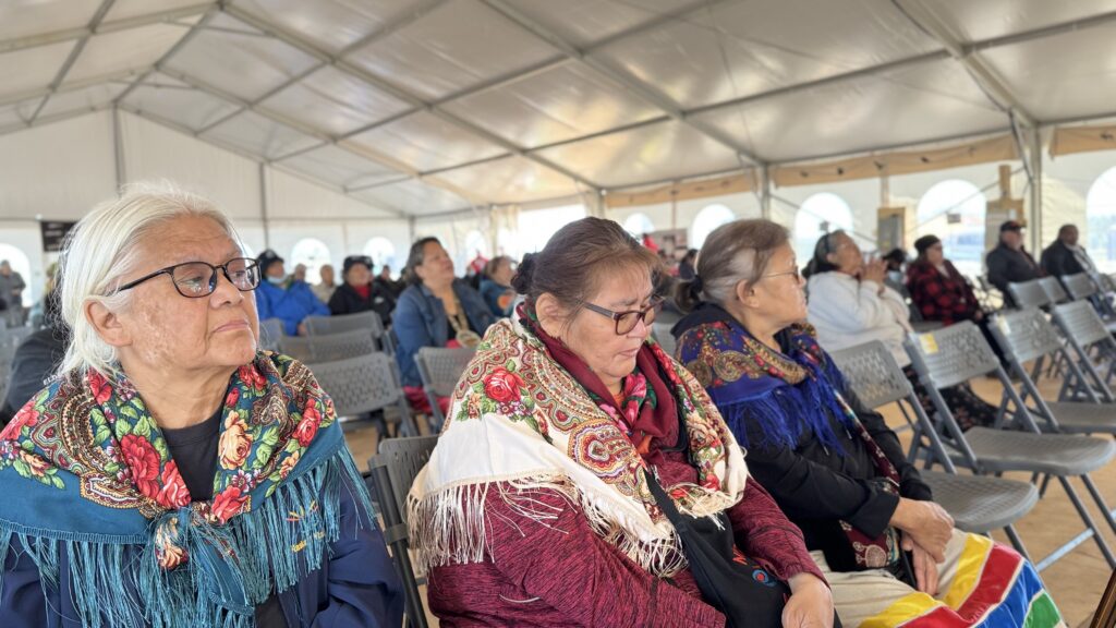 A group of elder Naskapi women, all wearing intricately patterned shawls, listens intently during a gathering. The expressions on their faces are thoughtful and reflective, as they absorb the stories and messages shared during the mass. Their colorful scarves and peaceful demeanor offer a rich tapestry of tradition, wisdom, and quiet strength, highlighting the community's respect for their elders and spiritual practices.