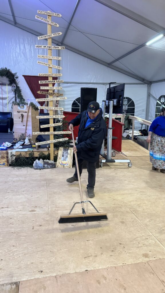 A man wearing a cap and jacket sweeps the wooden floor inside a large tent with a long broom. Behind him, there is a tall wooden signpost with distances to various indigenous communities written on it, such as "Matimekush 16 km" and "Sheshatshiu 473 km." The space is set up for an event, with equipment and decorations visible around the stage area.