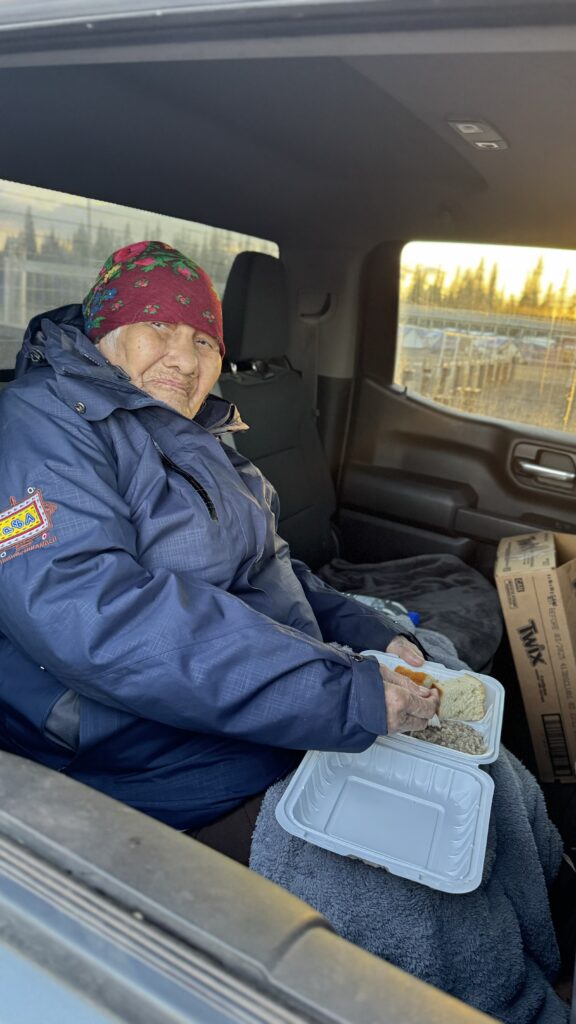 An elderly woman sits in the back seat of a car, holding a white to-go container with a meal of bread and other food. She wears a jacket with a colorful Naskapi patch on the sleeve and a red floral headscarf. Her expression is content, as she enjoys the meal in a cozy setting.