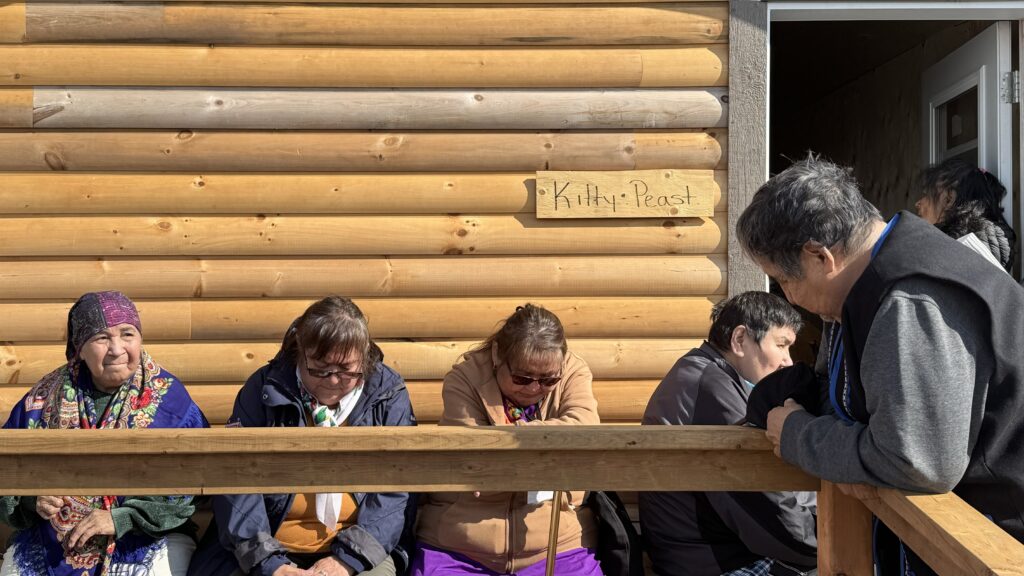 A group of elderly Naskapi people sit on a wooden porch outside a log cabin. Some are sitting quietly with their heads bowed, while a man stands by the door leaning on the railing. Above them, a small hand-made sign reads "Kitty Peast." The bright sunlight highlights the wood of the cabin and the gathering on the porch.