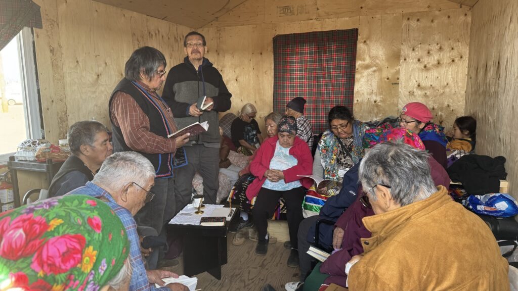 Inside a wooden cabin, a group of Naskapi people gather for a church service. Several elderly individuals sit on benches and chairs, holding hymn books or prayer guides. Two men stand reading from books, while the rest of the group listens or follows along in their seats. The room has simple wooden walls and a plaid fabric covers one of the windows.
