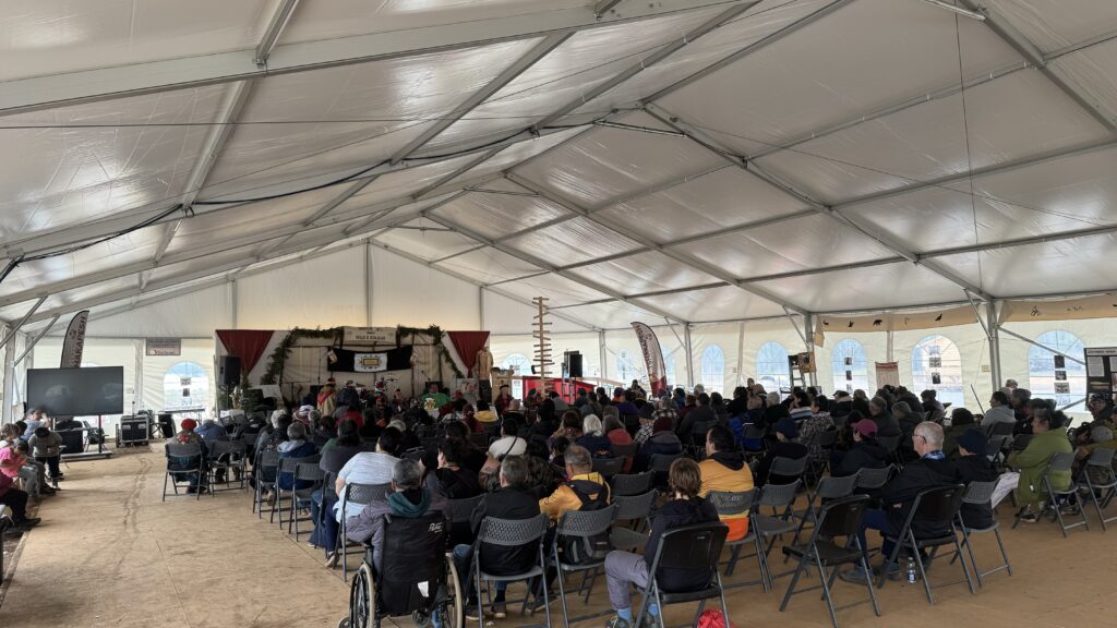 Inside a large tent filled with rows of chairs, a crowd of people attends a church service. At the front, there’s a decorated stage with a banner that reads "Kwe," alongside a podium. The service includes elements of indigenous culture, with banners and symbols visible on the walls.