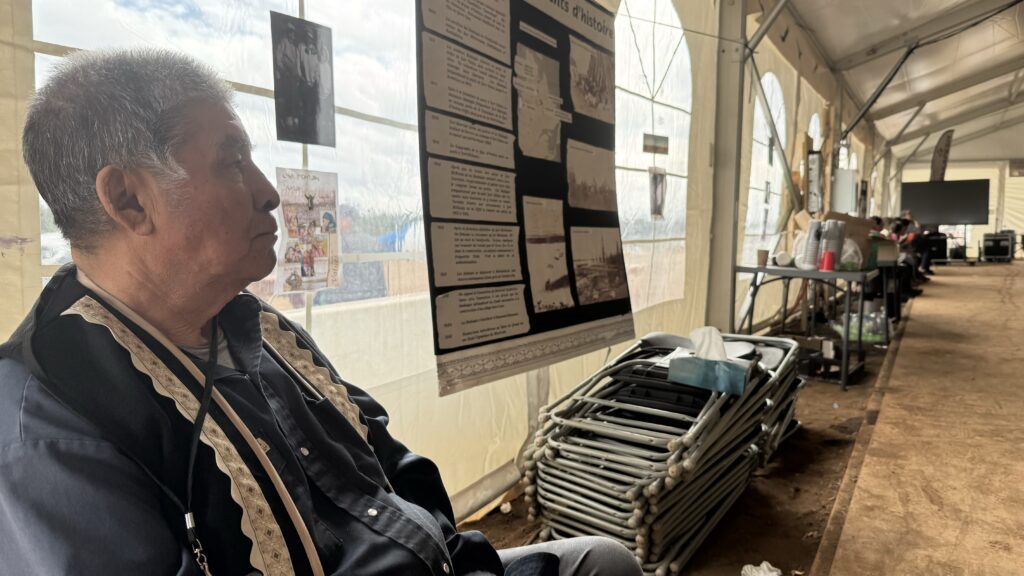 An elderly man in a traditional black jacket with beige trim sits inside a large tent, gazing thoughtfully at a display. Behind him, historical posters are mounted on the wall, and stacks of folded metal chairs rest nearby. The tent has a spacious, well-lit interior with other people in the background.