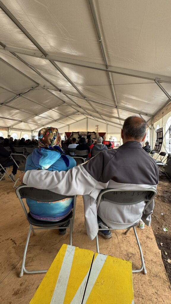 A man and a woman sit side by side inside the tent, viewed from behind. The man wraps his arm around the woman's shoulders, comforting her as they sit close together. The scene focuses on their intimate moment as they attend the service with others seated in the background.