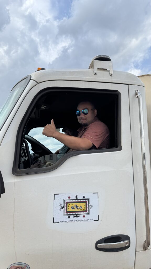 A man sitting in the driver's seat of a large white truck gives a thumbs-up through the window. The side of the truck displays a decal that reads "Naskapi Nation of Kawawachikamach." The cloudy sky provides a backdrop to the scene, highlighting the driver’s role in transporting supplies.