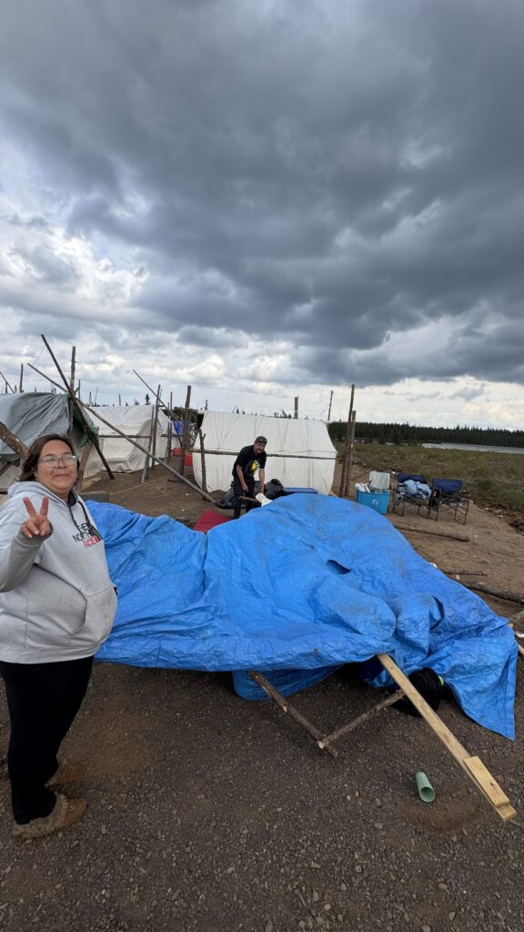 A woman stands on a gravel surface next to a collapsed tent covered in a blue tarp, giving a peace sign with her hand. In the background, another person is working near other tents made of wood and tarps, set up near a river. The sky is cloudy and dark, with strong winds affecting the camp setup.