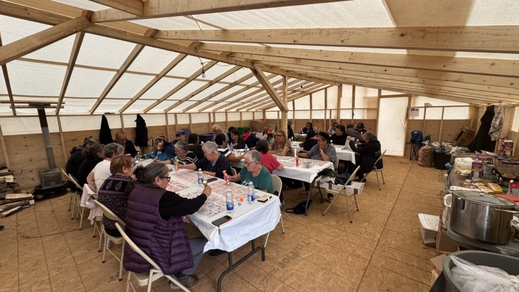 Inside a large, low-built kitchen tent, elders sit at long tables covered with bingo cards and markers. The room is filled with the sound of conversations and laughter as the group enjoys playing bingo, sipping tea, and coffee. The wooden frame of the tent is visible, and there is a wood stove with logs stacked nearby, contributing to the cozy, communal atmosphere.