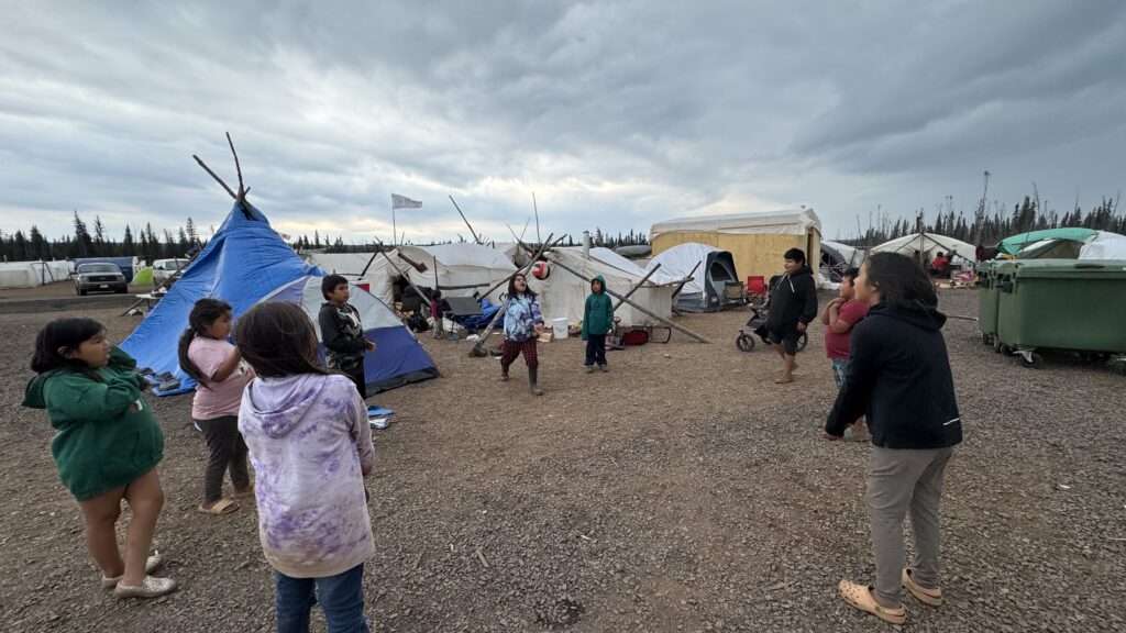 A group of children stands in a circle on a gravel surface, tossing a volleyball back and forth between tents. A blue teepee and various makeshift structures are visible in the background, with a cloudy sky overhead hinting at an approaching storm. The children seem undeterred by the weather, continuing their game.