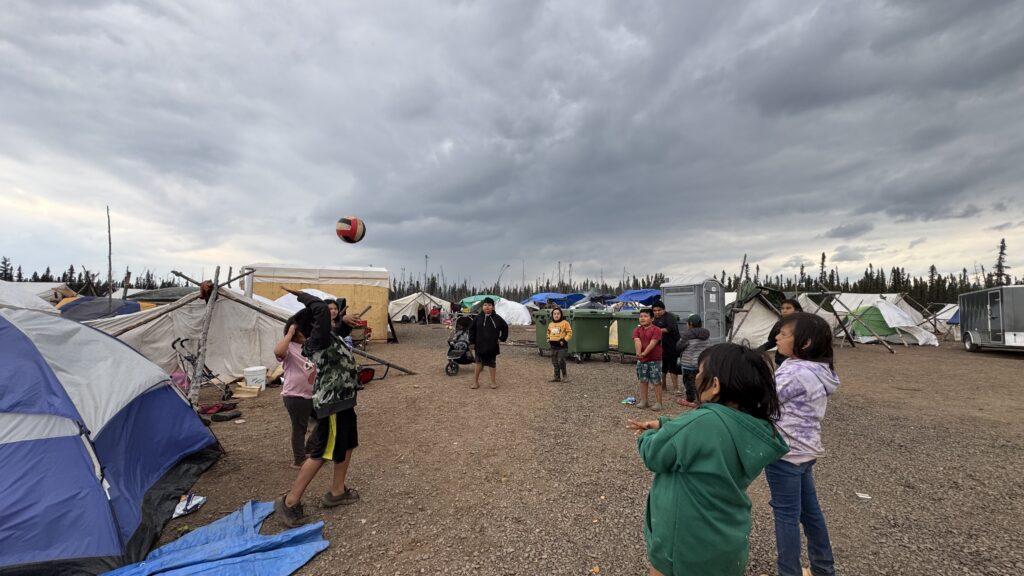 A wider view of the children playing volleyball at the camp, with more tents and a trailer visible in the background. The children are engaged in the game, with the volleyball suspended mid-air. The cloudy, stormy sky looms above the camp, but the kids remain focused on their game.