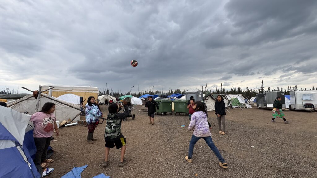 A close-up of the volleyball in mid-air as the children continue playing in the open area between tents. The game takes place in a rustic campsite, with the storm clouds in the background adding a dramatic touch to the scene.