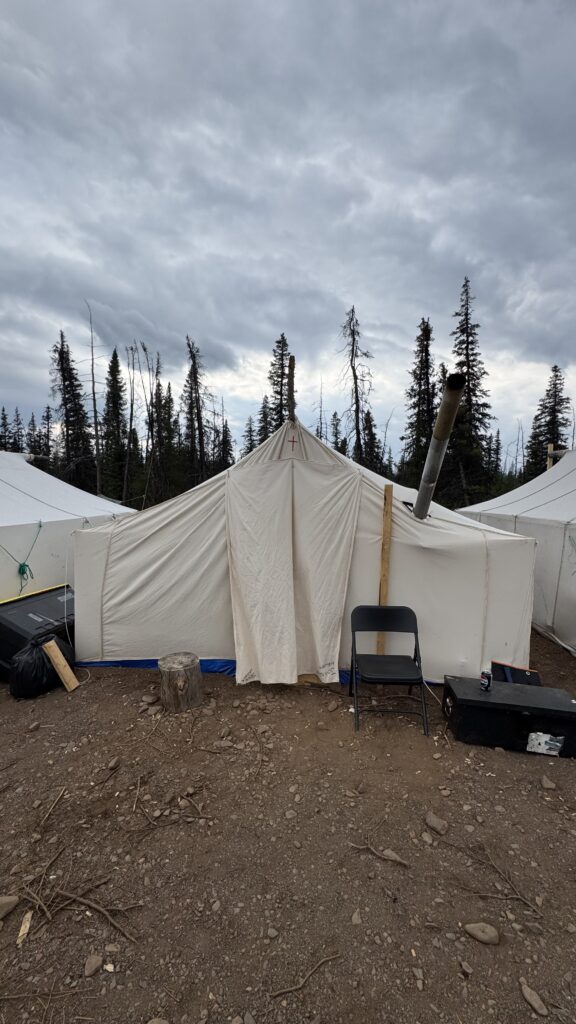 A large white canvas tent with a small cross at the top sits on rocky ground, with a metal stovepipe protruding from one side. A black chair and a log stump are positioned outside the entrance, and the tent is surrounded by sparse trees and cloudy skies in the background, giving a rustic, camp-like feel.