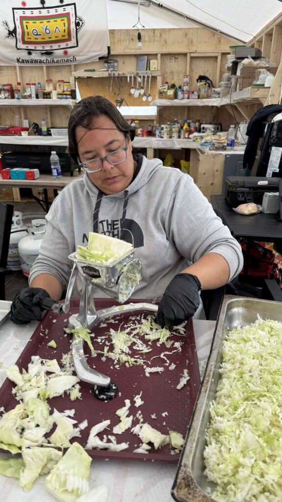 A woman wearing a gray hoodie is seated in a kitchen tent, manually shredding cabbage using a hand-cranked shredder. She is focused on her task, with gloves on, and the shredded cabbage falls onto a tray in front of her. The tent’s shelves behind her are stocked with supplies, and a banner reading "Kawawachikamach" hangs above.