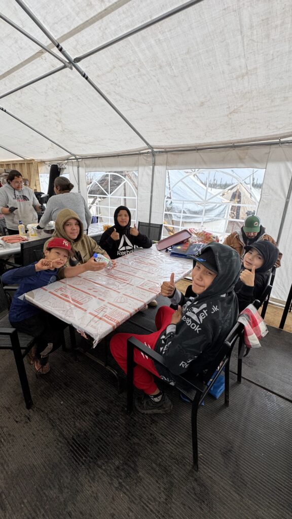 A group of children sits at a long table covered with a plastic tablecloth inside a kitchen tent, sheltering from the rain. They give thumbs-up to the camera, wearing hoodies and caps. Despite the rainy weather, they appear cheerful, with food and snacks in the background, while the camp outside can be seen through the tent windows.