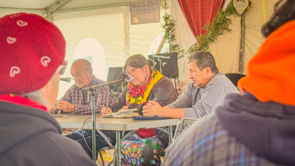 A group of three people seated at a table under a large tent, holding a religious mass. A man in a plaid shirt reads from a book into a microphone, while the woman next to him, wearing a vibrant yellow scarf and floral skirt, also reads attentively. The third man, seated beside her, has his hands folded on the table. In the foreground, attendees are listening, some wearing bright hats and scarves. The tent is decorated with greenery, and the scene conveys a sense of community and spiritual reflection.