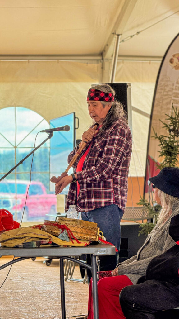 An elder stands with a ceremonial tobacco pipe during a traditional tobacco pipe ceremony, an important cultural ritual. They are dressed in a checkered shirt and headband, and nearby, another elder assists with the ritual preparations. Sage burns softly on the table in front of them.