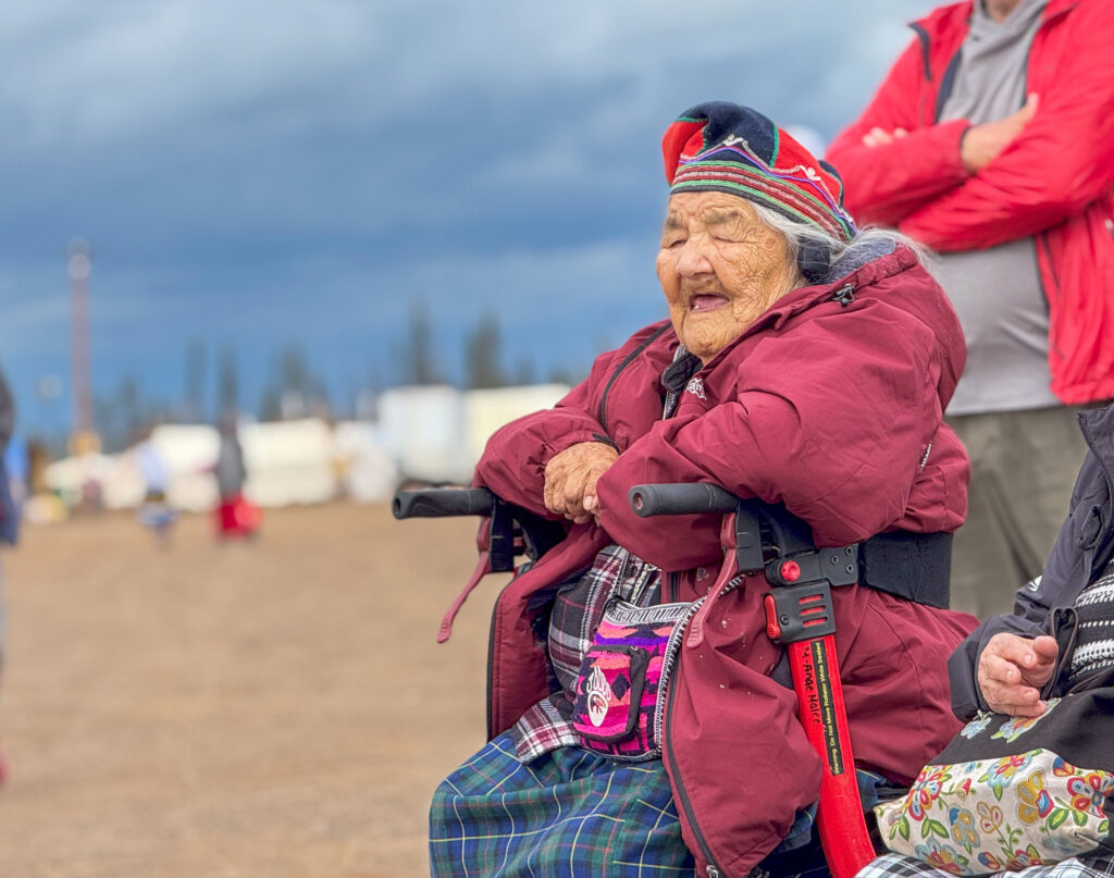 Walking Out Ceremony group (elder woman in red coat): An elder  woman, wearing a vibrant red coat and traditional cap, sits comfortably in a mobility scooter. She smiles warmly, clearly enjoying the ceremony. Behind her, a man in a red jacket stands with his arms crossed, as a dark sky looms in the distance.