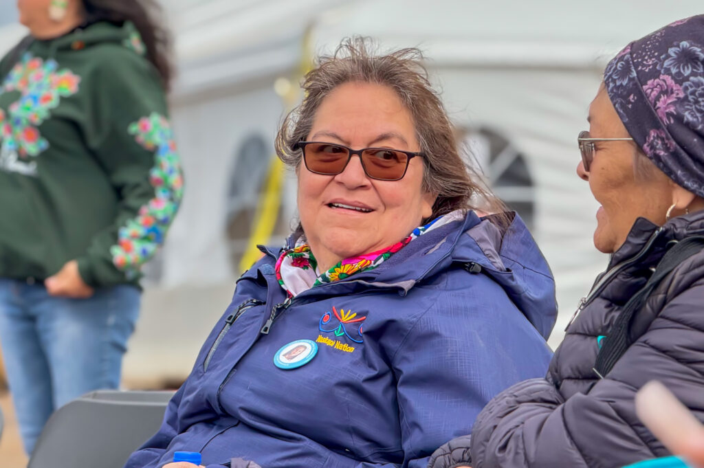 Walking Out Ceremony group (woman in blue jacket): A Naskapi woman in a blue jacket with the "Naskapi Nation" logo smiles and chats with another woman during the Walking Out ceremony. She’s wearing a colorful scarf, and a crowd can be seen mingling behind her.