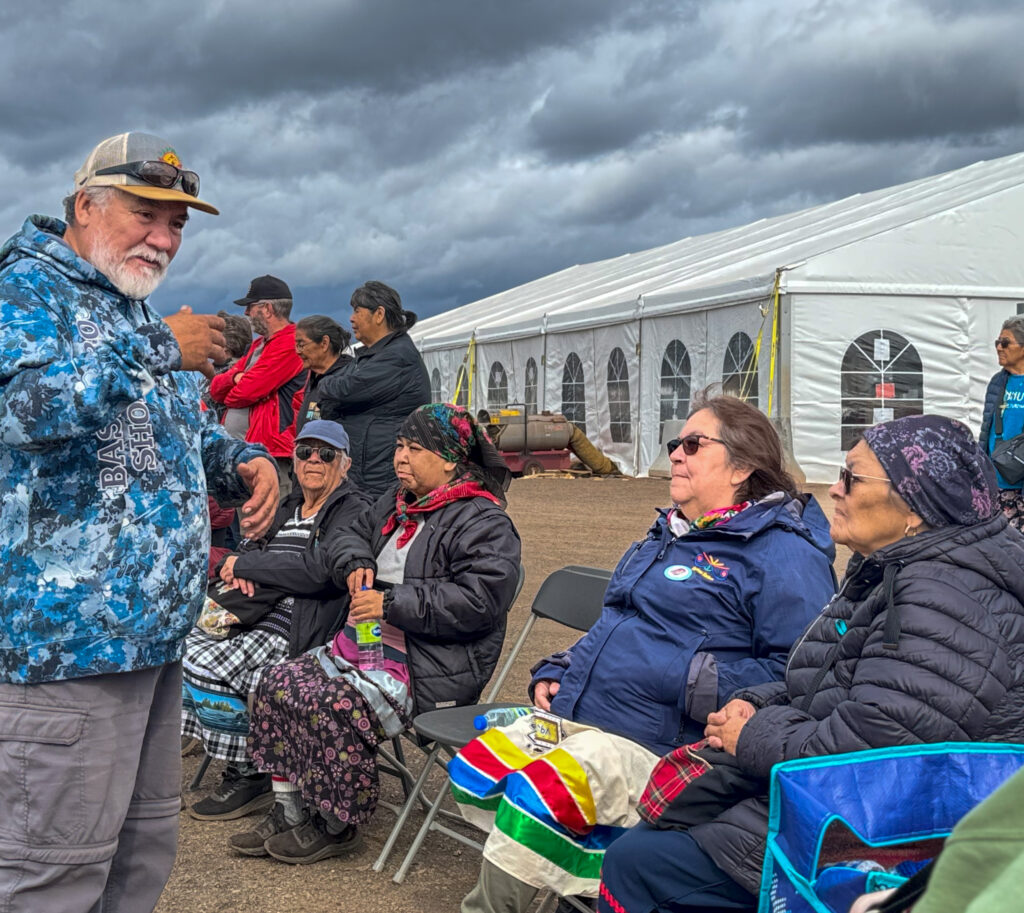 A group of older adults sit outdoors near a large white tent, bundled up in jackets and colorful blankets. A man in a blue camo hoodie speaks animatedly to the seated group, which appears to be listening attentively. The sky is overcast, and the gathering conveys a sense of community amidst traditional cultural elements.