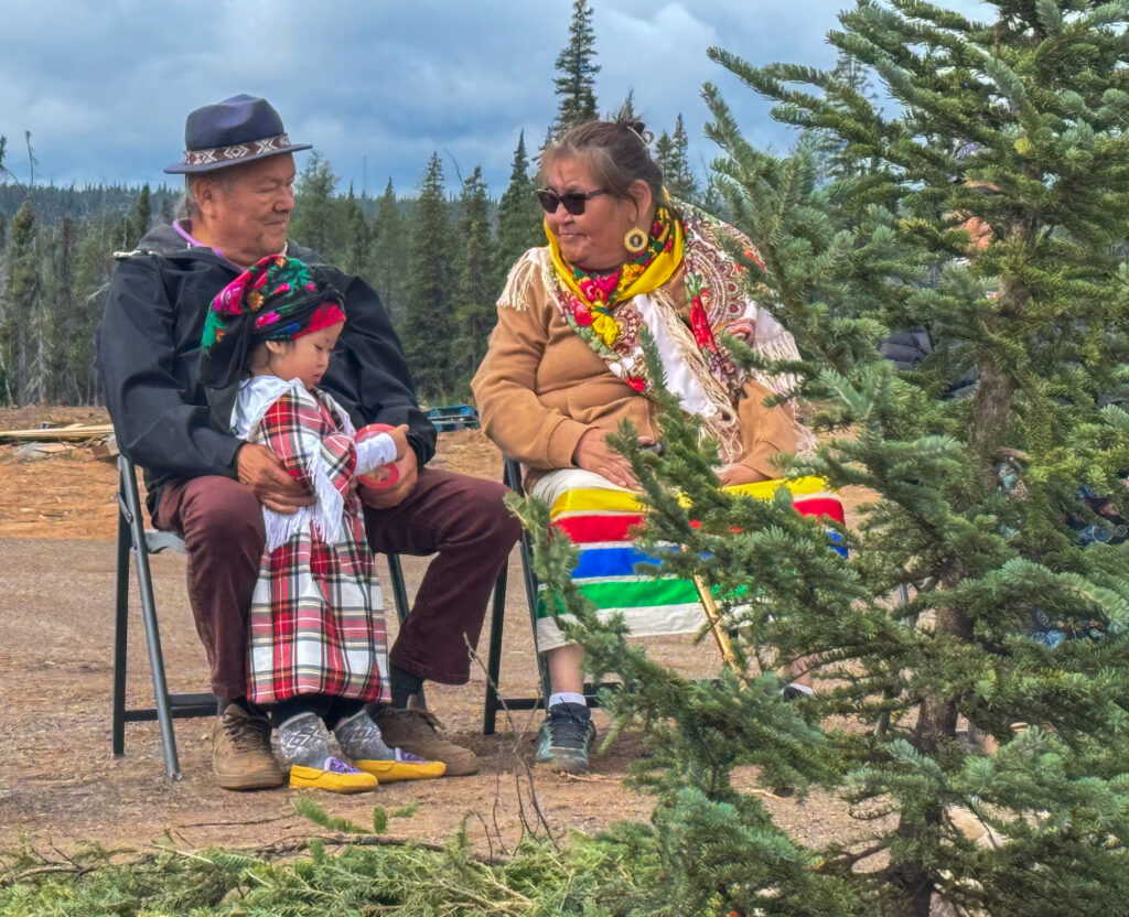 An older man, a young child in traditional plaid clothing, and an older woman sit together outdoors. The child sits on the man’s lap, holding a cup, while the woman, draped in a colorful shawl and blanket, smiles. The backdrop features evergreen trees and a cloudy sky, with a calm outdoor setting.