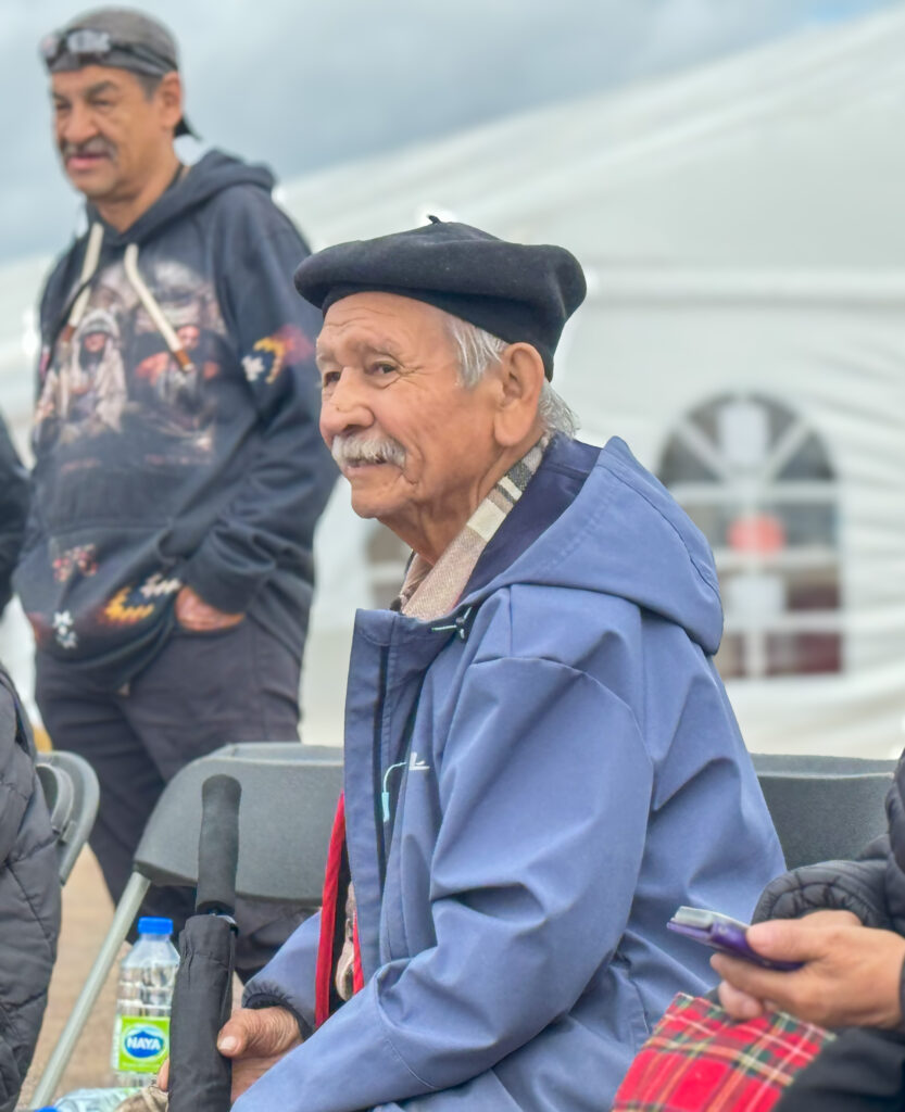 A seated elderly man wearing a blue jacket and black beret looks off to the side, smiling, while holding an umbrella. Behind him, another man stands in the background, dressed in a hoodie. A large white tent and an overcast sky fill the scene, creating an atmosphere of community gathering.