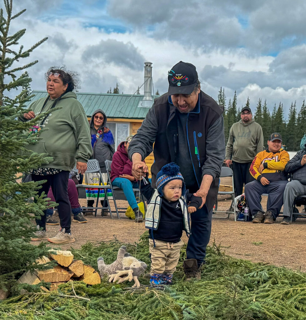 A man assists a young child, who is learning to walk on a ground covered in green fir branches. The man, dressed in a black vest and baseball cap, holds the child’s hands as they take steps. The child, bundled in a blue knit hat, stands near some wood and greenery, with seated people and cabins in the background.