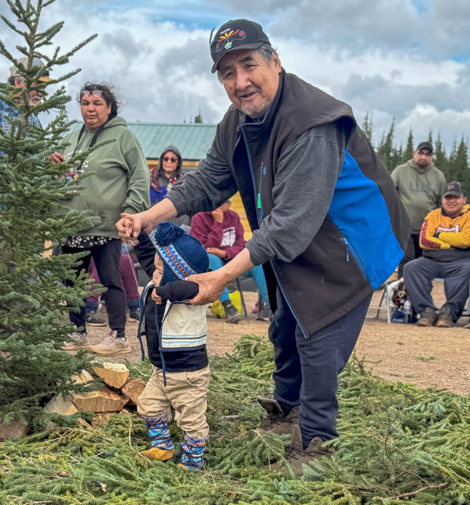 The same man continues to help the young child walk outdoors. The child, wearing a winter hat and boots, takes cautious steps across a patch of fir branches, as onlookers in colorful clothing watch from behind. The setting features an outdoor gathering with a mix of cultural and natural elements.