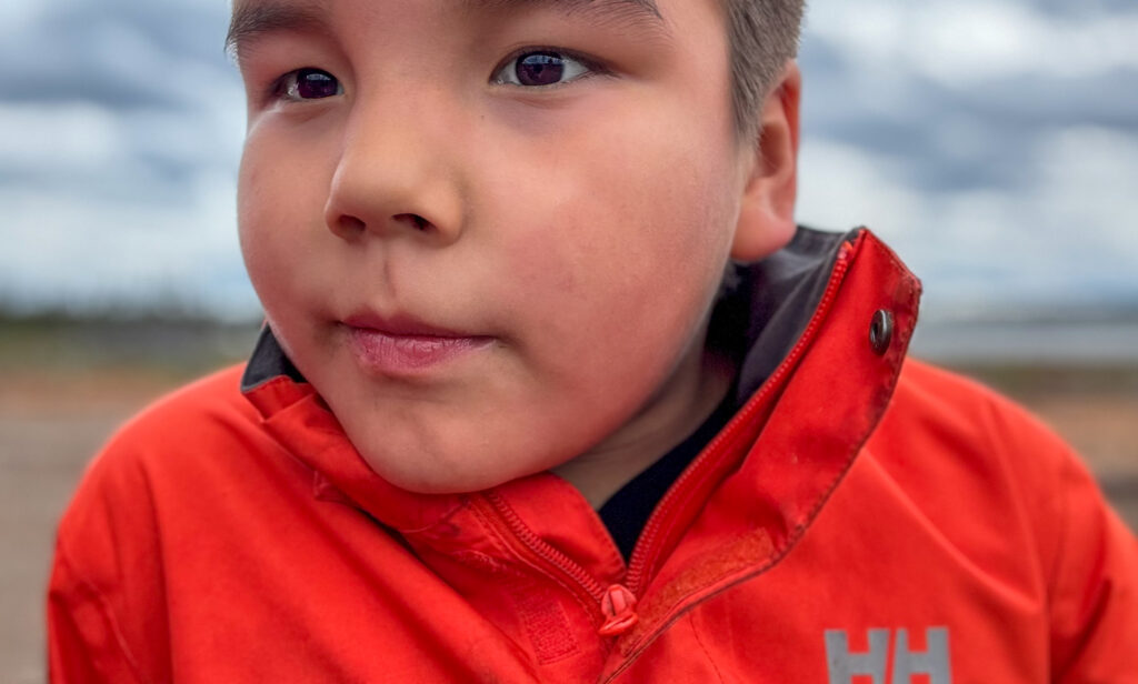 A close-up of a young boy, Cohen, wearing a bright red jacket, gazing curiously off to the side. His face is calm, and his eyes reflect natural light. The blurred background hints at an outdoor setting with soft, overcast skies.