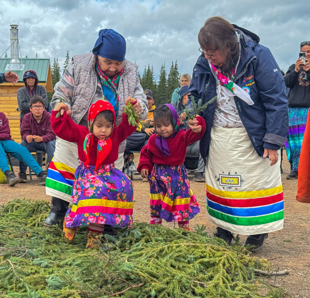 Two older women, each holding a young girl by the hands, guide them as they walk on a bed of pine branches. The young girls, dressed in bright red sweaters and colorful skirts, hold small evergreen sprigs. A crowd of people sits in the background, watching the ceremony unfold in an outdoor setting.