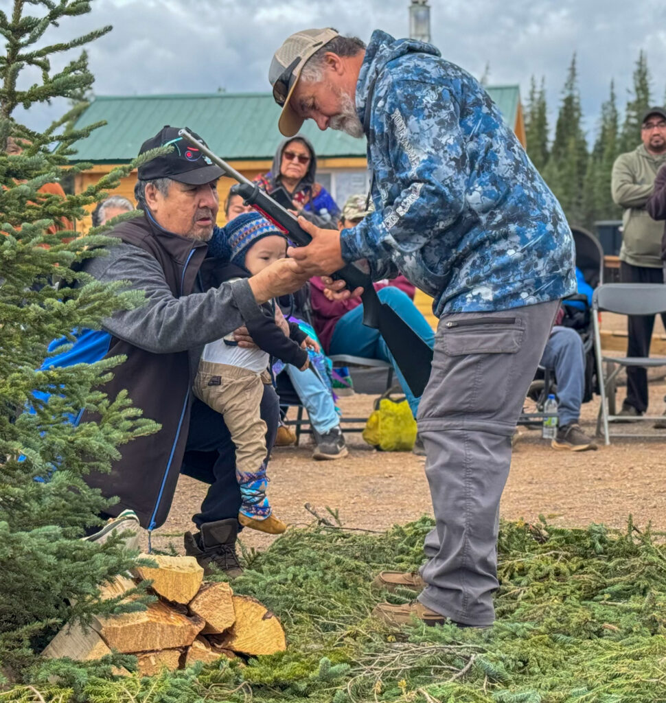 A man helps a young boy, who is being held by another man, hold a rifle. The scene takes place near a pile of wood and pine branches, with people watching the ceremony from behind. The boy is bundled in a blue hat and winter boots, while the men guiding him wear casual outdoor clothing.