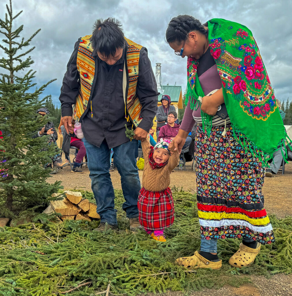 A smiling toddler in a red plaid skirt and brown sweater is supported by two adults holding her hands as she steps on a bed of pine branches. The adults, dressed in colorful clothing with traditional designs, stand next to a small evergreen tree as part of the ceremonial scene.