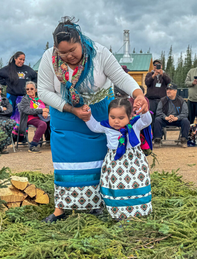 A woman, dressed in a white and blue traditional outfit, gently holds the hands of a young girl in matching attire as they walk on a bed of pine branches. The girl clutches an evergreen sprig in one hand, while onlookers behind them, including elders and other participants, observe the event in an outdoor setting.