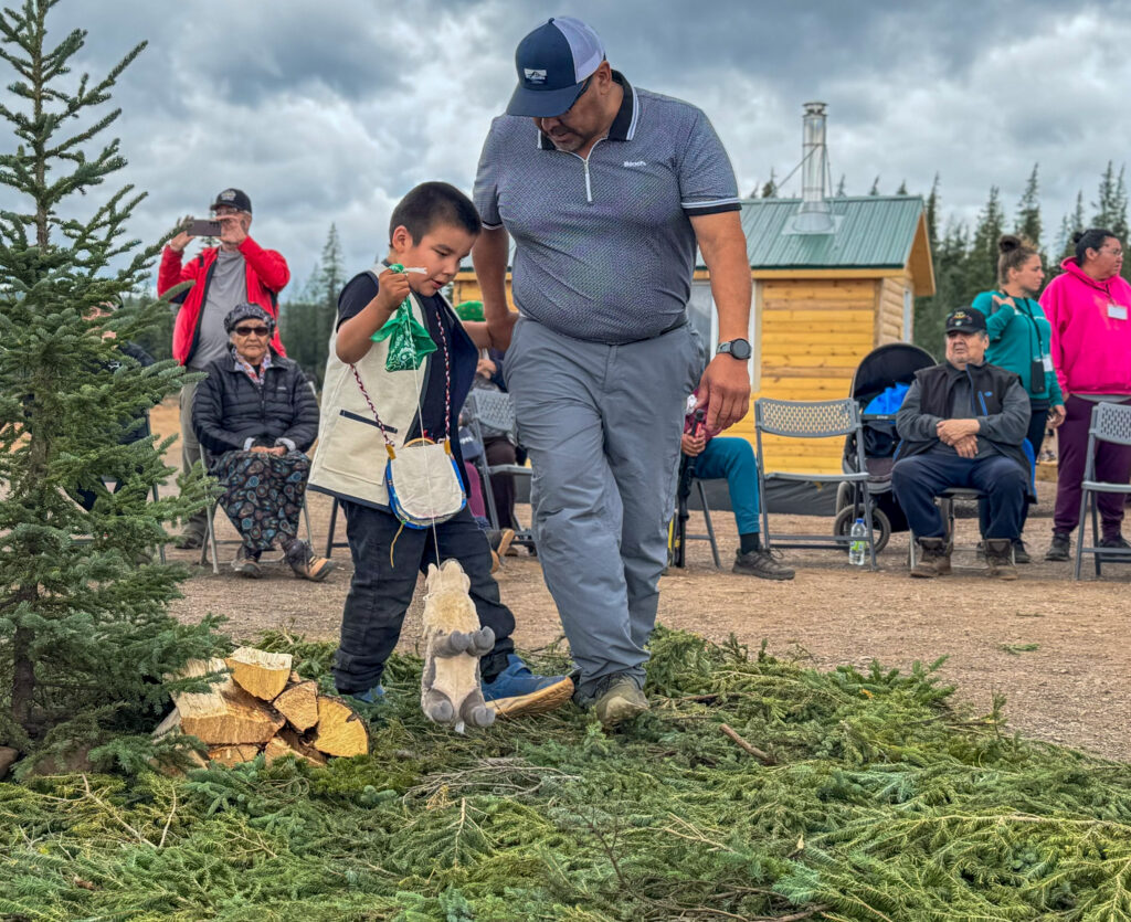 The same man and young boy continue their ceremonial walk over pine boughs. The boy holds a green cloth and a small stuffed animal representing a caribou as they pass a pile of chopped wood. People in the background, some taking photos, observe the significant moment under cloudy skies.
