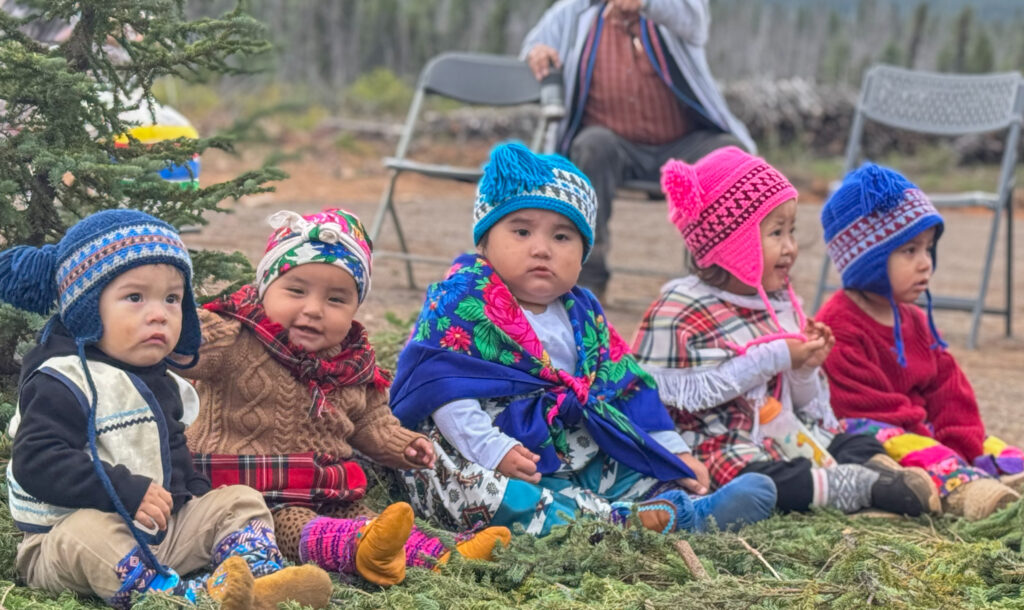 A group of young children, seated in a line, wearing colorful knit hats, traditional outfits, and cozy boots, look ahead with curious expressions. They sit on a bed of pine branches outdoors, next to a small evergreen tree. The background is blurred, with some chairs and an elder watching the event.