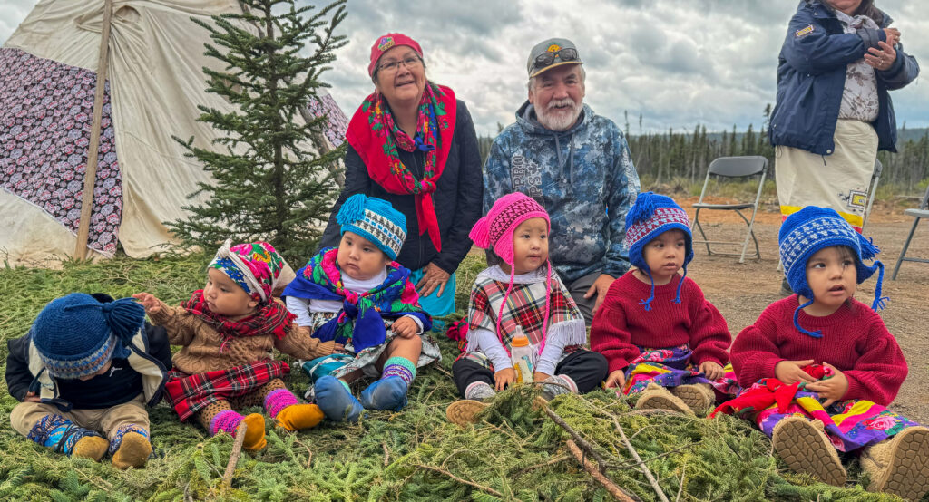 Elders and two adults sit behind a group of young children who are seated on pine branches. The children are dressed in vibrant, traditional clothing with colorful hats, and the elders are smiling, observing the scene. A large tipi structure and an evergreen tree are visible in the background.