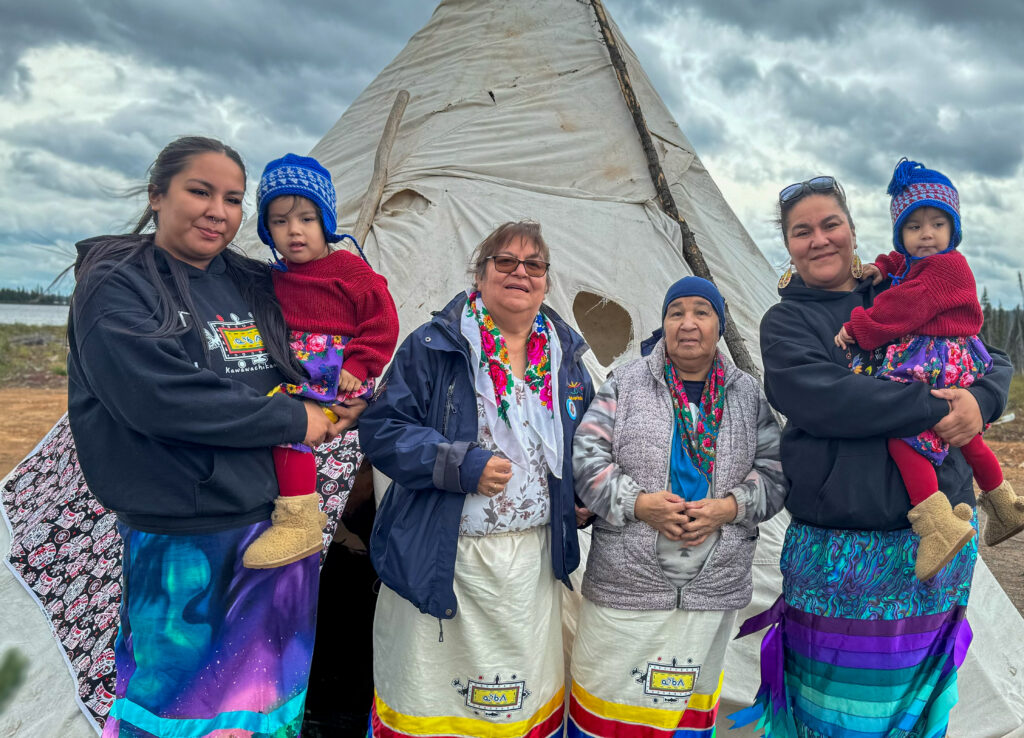 A group of three women and two young children pose outside a tipi. The children wear matching blue knit hats and red sweaters while being held by two of the women. The adults are dressed in traditional skirts with colorful designs, smiling as they celebrate the ceremony together.