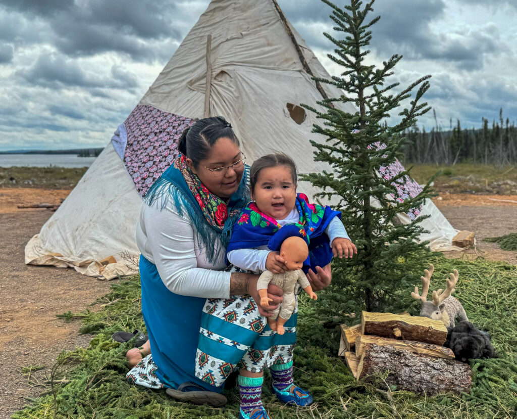 A woman helps a young child, who is dressed in traditional clothing, kneel beside a small evergreen tree during the ceremony. The child holds a doll, and a stuffed caribou toy rests nearby on a pile of logs, symbolizing elements of traditional Naskapi life. Behind them, a tipi stands under cloudy skies.