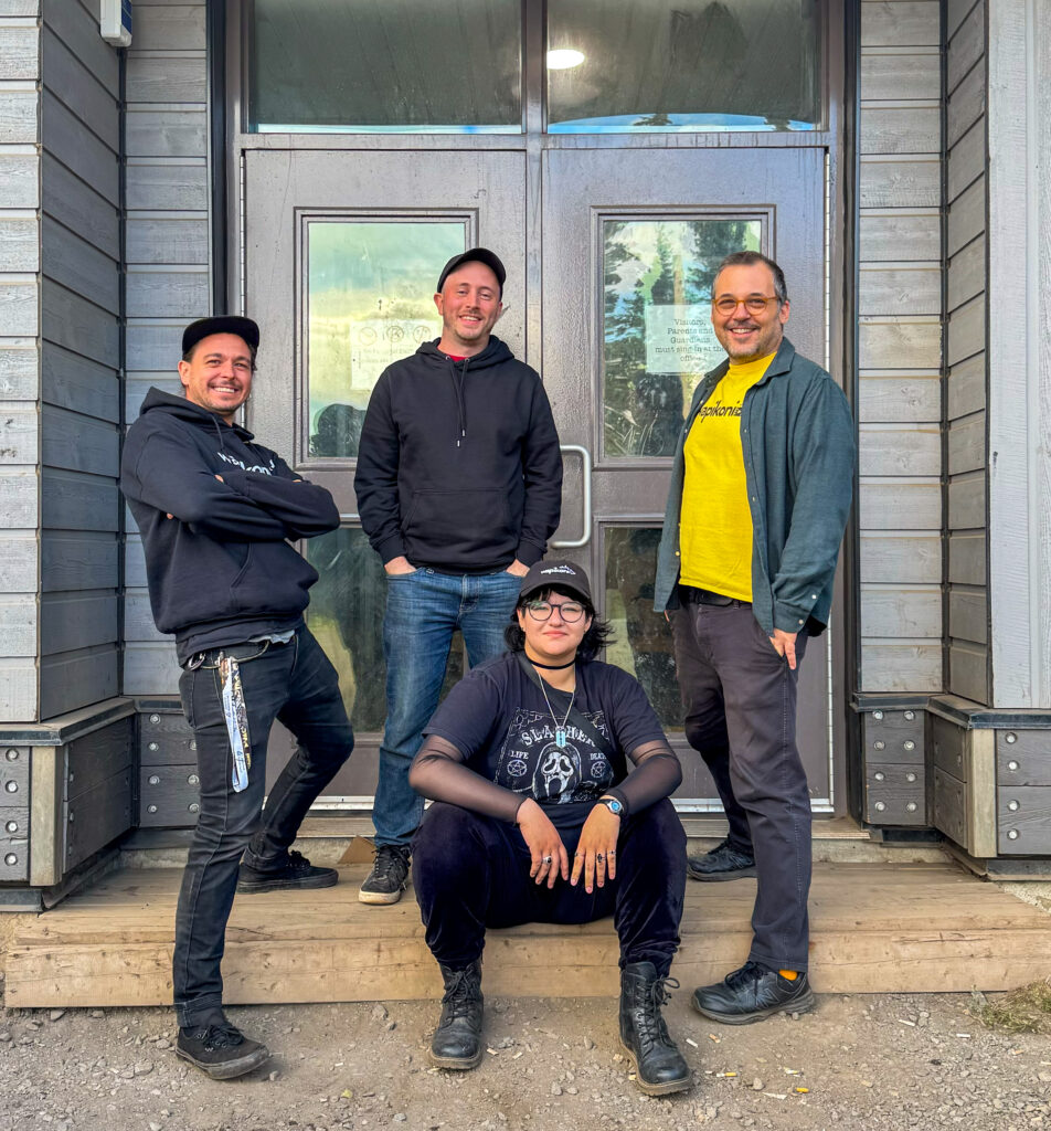 Four people stand in front of a building's door, smiling for the camera. Three of them are standing while one person sits on the steps in front. They are dressed casually, wearing hoodies, jeans, and T-shirts. The backdrop is a modern building with a glass door, and the group appears relaxed and friendly. This is part of the Wapikoni team, collaborating with the Naskapi Nation to record and produce music and videos.