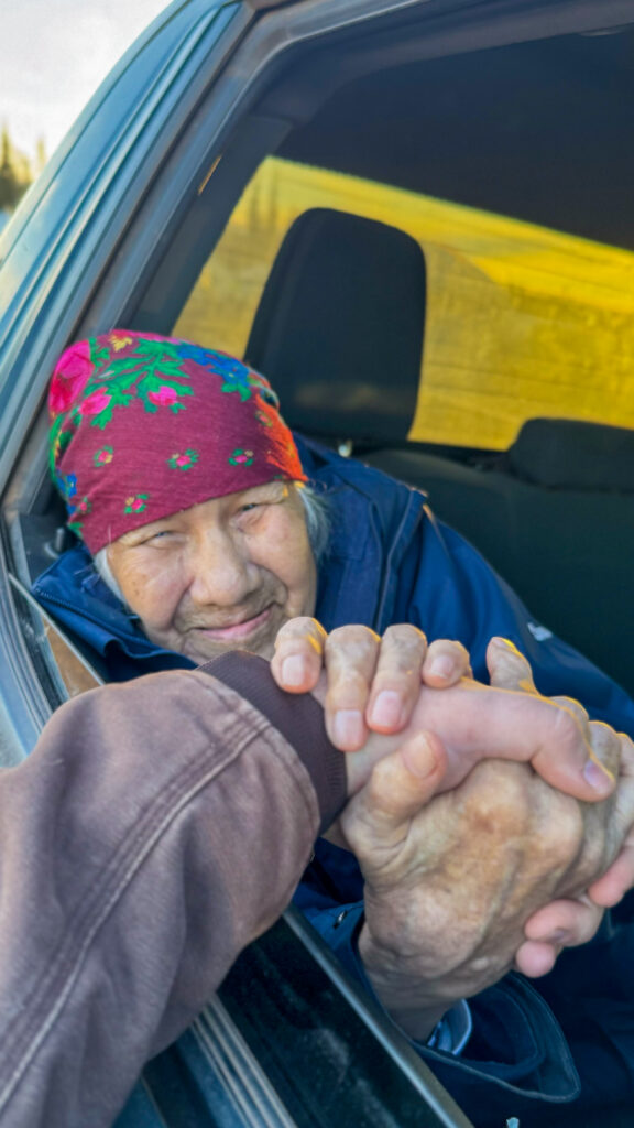 An elderly woman, wearing a colorful red headscarf with floral designs, reaches out from a car window, holding someone’s hand firmly. Her expression is warm and affectionate as she leans slightly out of the car. The image captures a tender, intimate moment between the two people.